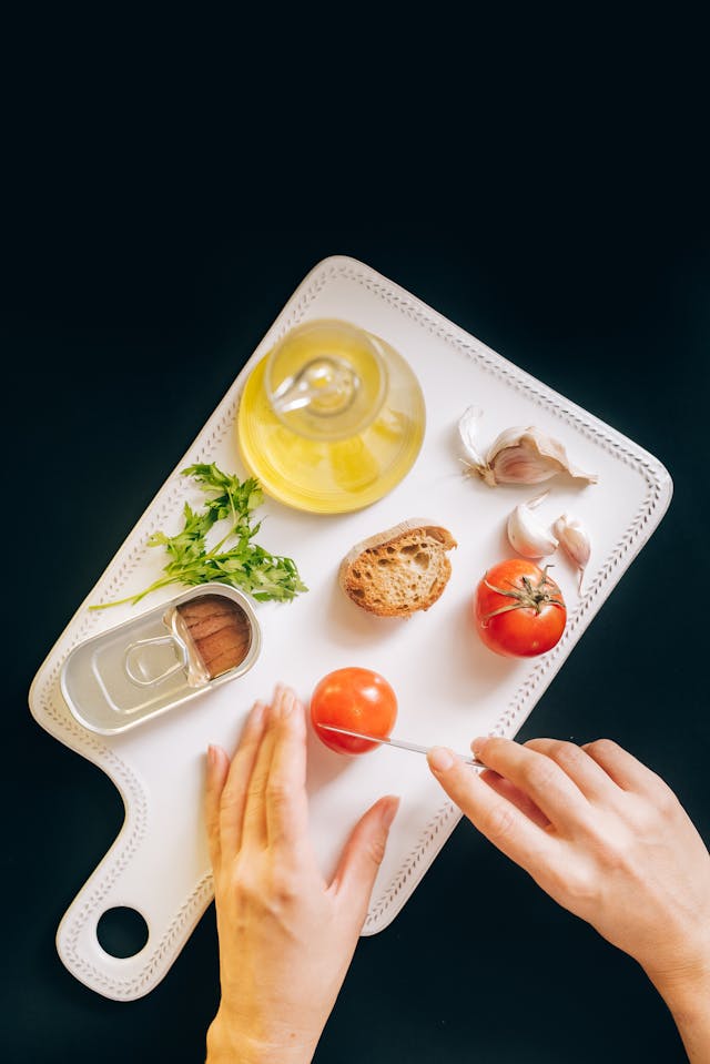 Low carb keto meal preparation: fresh tomatoes, garlic, olive oil and bread on a cutting board with hands slicing ingredients for a Mediterranean-style lunch recipe