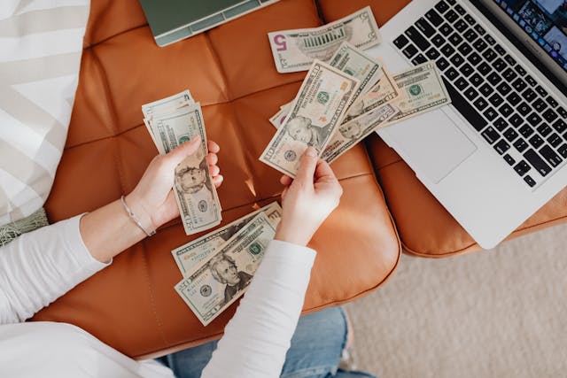 A woman counting money on a leather couch with a laptop nearby, symbolizing the cost-saving benefits of the Metabolic Reset Diet for budget-friendly fat loss.