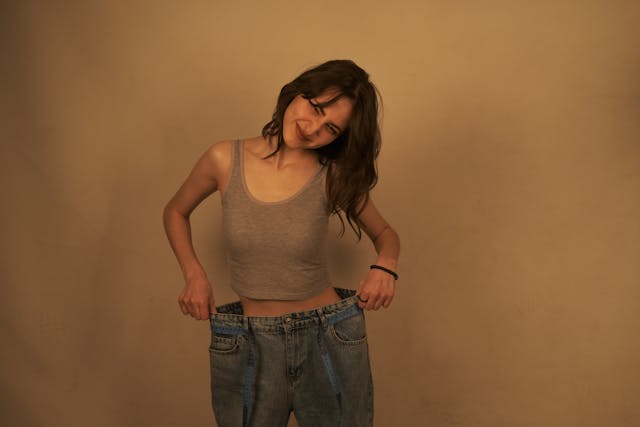 A smiling woman proudly stands in oversized jeans, showcasing significant weight loss and a transformed figure achieved with the Metabolic Reset Diet.