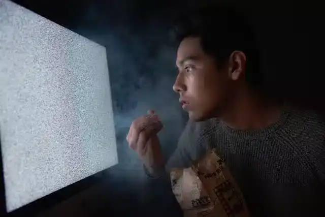 Shift worker sitting in dark room in front of large monitor holding a food package, illustrating circadian fasting challenges during night shifts